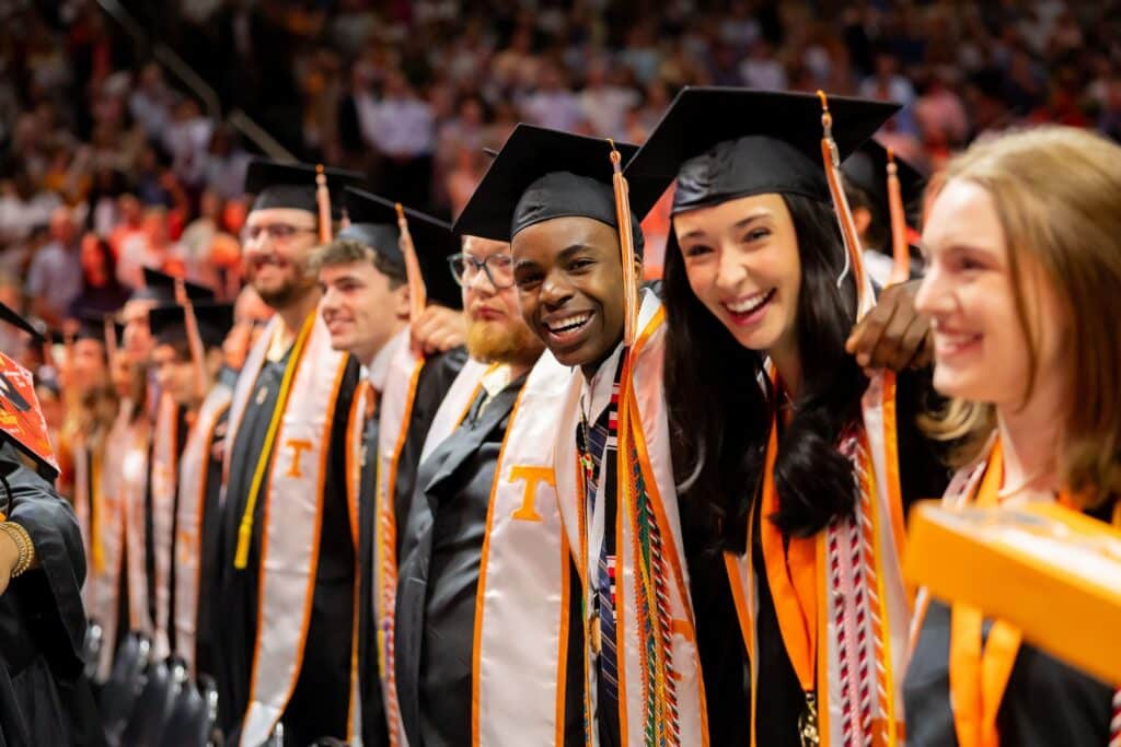 A group of graduates in black caps and gowns with orange and white sashes smile broadly during a commencement ceremony.